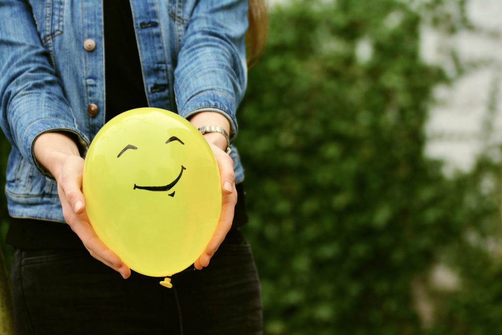 hands holding a yellow balloon with smiley on to invite you to Bring Joy to Another